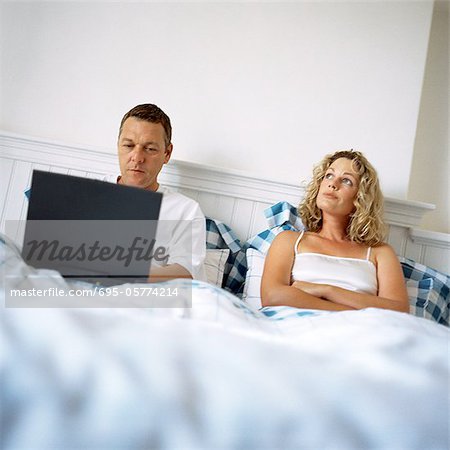 Couple in bed, man using laptop computer, woman with arms crossed