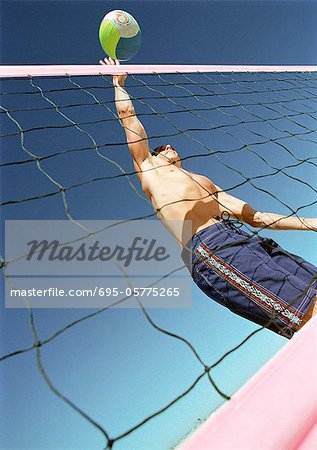 Young man playing beach volleyball, hitting ball over net, low angle view.