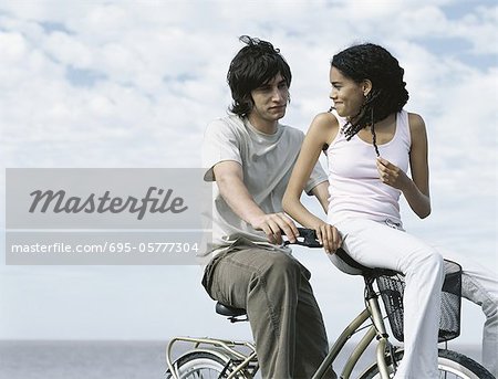 Young man sitting on bicycle, young woman sitting on bicycle basket looking over shoulder at young man