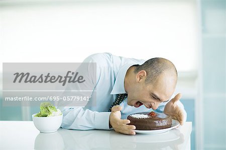 Man sitting at table with chocolate cake and bowl of lettuce, leaning toward cake with mouth wide open