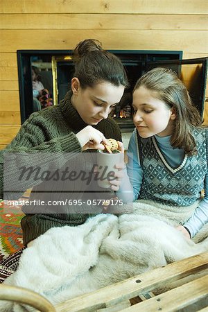 Two teenage girls sitting by fireplace, sharing snack