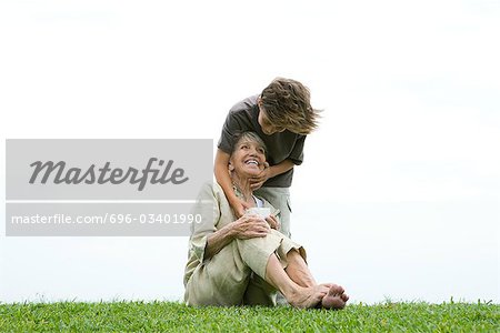 Senior woman sitting on grass, grandson standing behind her with hand on her face, giving her gift