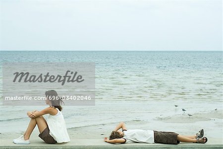 Two young friends looking at ocean, one sitting, the other lying on back