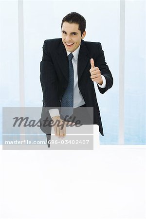 Businessman leaning on back of chair, giving thumbs up to camera