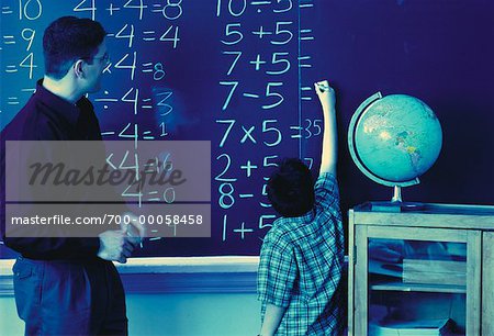 Teacher Watching Boy Writing On Blackboard in Classroom