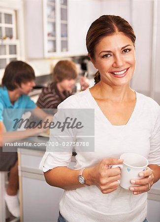 Mother in Kitchen With Cup of Coffee, Sons Eating Breakfast in the Background