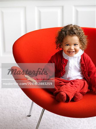 Little Girl in White and Red Outfit Sitting on a Red Chair in a White Room