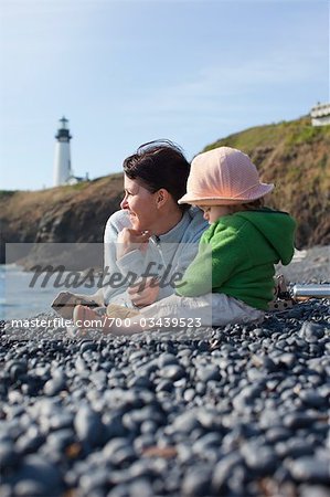 Mother and Baby Daughter Sitting on Beach, with Yaquina Head Lighthouse in the Distance, near Newport, Oregon, USA