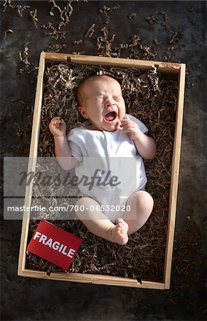yawning newborn baby girl wearing white undershirt onesie in a shipping box labeled as fragile with packing paper