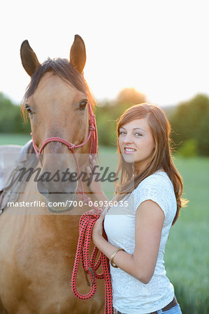 Young attractive woman standing beside a horse on a meadow by sunset, Germany