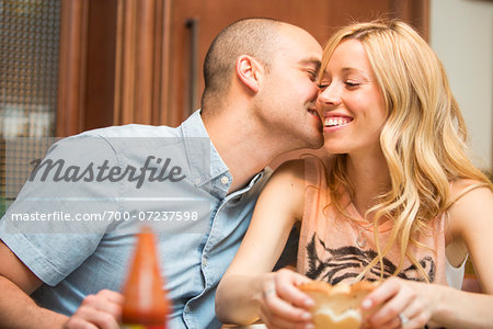 Young man kissing young woman on cheek, sitting and eating in home, Canada