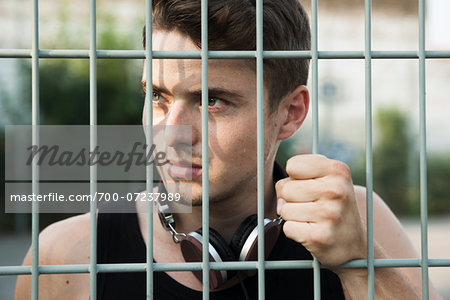 Close-up portrait of young man outdoors, standing behind steel fence with headphones around neck, Germany