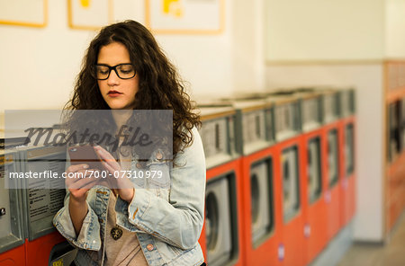 Teenage girl standing next to dryers in laundromat, using smart phone, Germany