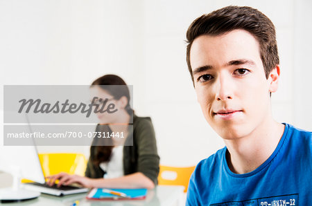 Teenage boy looking at camera, with teenage girl working on project using computer in background, studio shot
