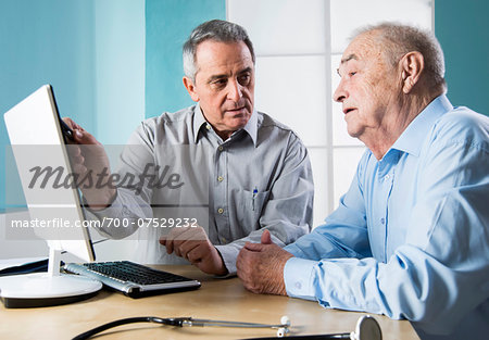 Senior, male doctor conferring with senior, male patient, using desktop computer in office, Germany