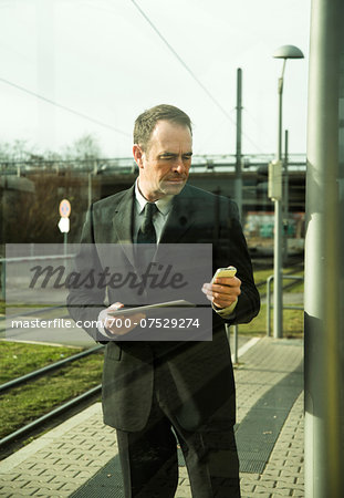 Businessman holding tablet computer and looking at cell phome, standing at train station outdoors, Mannheim, Germany