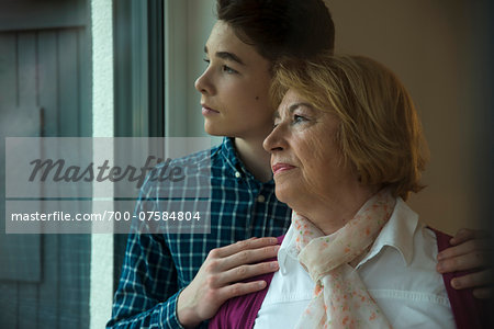 Close-up portrait of teenage boy with grandmother looking out window at home, Germany