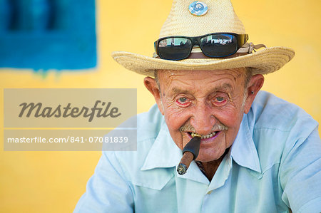 Portrait of old man wearing straw hat and smoking cigar, posing against a yellow wall for tourist pesos, Trinidad, Cuba, West Indies, Central America