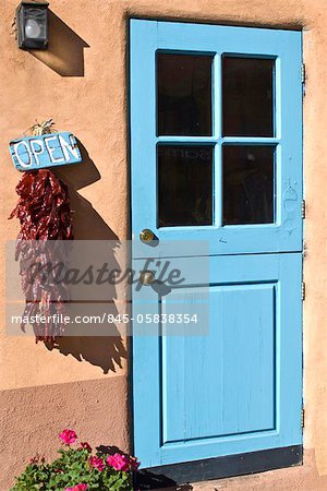 A turquoise-painted wooden stable-style door in an adobe wall, with a chilli ristra and a painted open sign, on Canyon Road, Santa, Fe, New Mexico.