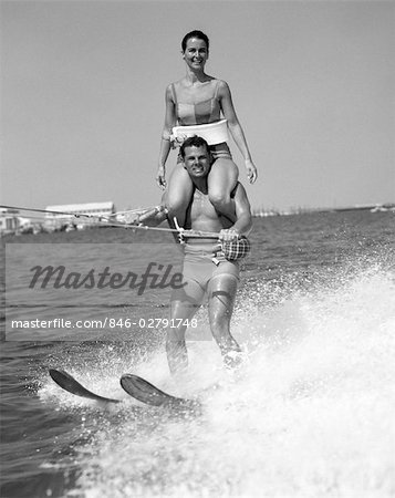 1960s MAN WATER SKIING WITH WOMAN IN BATHING SUIT RIDING ON SHOULDERS