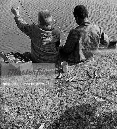 1960s BACK VIEW OF BLACK BOY AND WHITE BOY FISHING IN WATER WITH CAN OF WORMS & TACKLE BOX SHARED TOGETHER