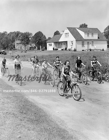 1950s GROUP OF 8 KIDS BOYS & GIRLS RIDE BICYCLES ON COUNTRY RURAL ROAD LANE FUN HOUSE IN BACKGROUND