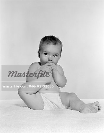 1950s BABY SEATED ON CARPET BRINGING FOOT TO MOUTH
