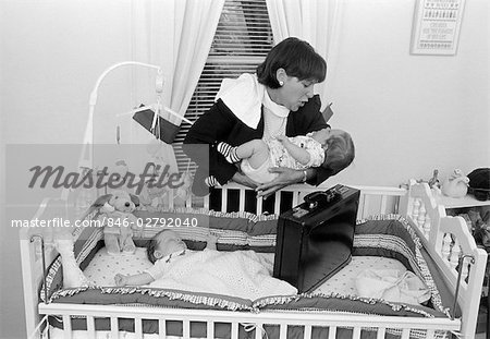 1980s WORKING MOTHER STANDING OVER CRIB HOLDING ONE BABY WHILE OTHER SLEEPS BRIEFCASE IN CRIB INDOOR