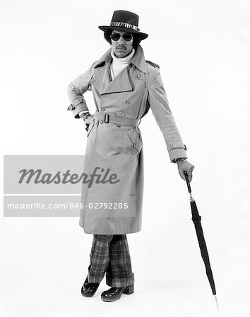 1970s PORTRAIT OF AFRICAN AMERICAN MAN DRESSED IN TRENCH COAT AND HAT LEANING ON UMBRELLA LOOKING AT CAMERA