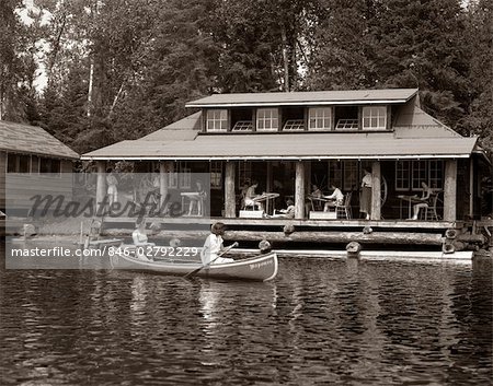 1950s TWO WOMEN PADDLING IN CANOE BY CABIN IN LAKE ALGONQUIN PARK CANADA SUMMER CAMP RUSTIC LOG CABIN