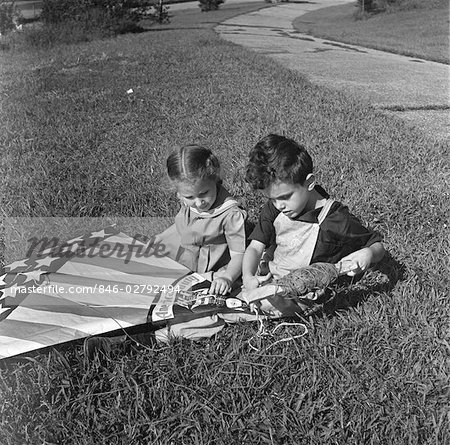 1940s GIRL AND BOY SITTING IN GRASS LAWN HOLDING AMERICAN FLAG STYLE KITE