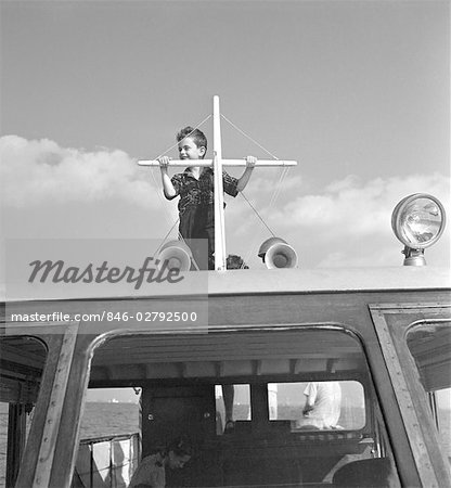 1940s BOY STANDING ON TOP OF MOTOR BOAT COCKPIT HOLDING ON TO SHORT MAST