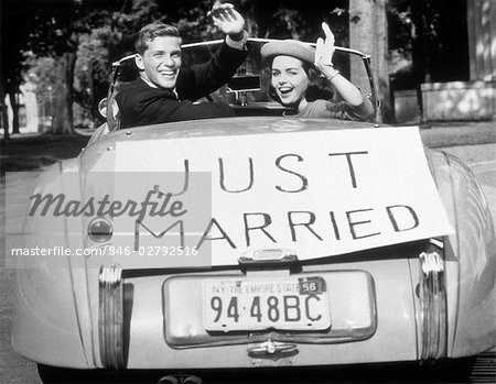 1950s NEWLYWED YOUNG COUPLE MAN WOMAN IN CONVERTIBLE SPORTS CAR WITH JUST MARRIED SIGN WAVING LOOKING AT CAMERA