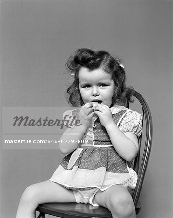 1930s TODDLER GIRL SIT IN WOODEN CHAIR POLKA DOT DRESS SHOWING UNDERPANTS EATING A COOKIE WITH BOTH HANDS VINTAGE