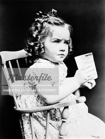 1930s CHILD GIRL SITTING IN HIGH CHAIR HOLDING GLASS OF MILK SERIOUS LOOK BOW IN HAIR BALONEY CURLS PRINT DRESS TODDLER