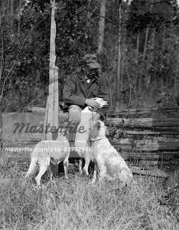 1920s ELDERLY MAN SITTING WITH RIFLE RESTING AGAINST FENCE PETTING ONE OF THREE HUNTING DOGS