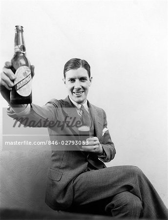 1930s MAN HOLDING UP HUGE DISTORTED BEER BOTTLE IN ONE HAND AND A REGULAR SIZE GLASS OF BEER IN OTHER  SMILING LOOKING AT CAMERA