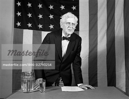 1940s ELDERLY MAN STANDING AT DESK GIVING SPEECH WITH 48-STAR AMERICAN FLAG IN BACKGROUND