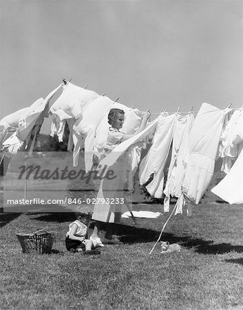 1930s WOMAN IN WHITE COTTON DRESS HANGING A FULL LINE OF CLOTHES IN THE BREEZE WITH A LITTLE BOY & CAT AT HER FEET