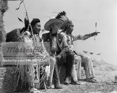 1940s PAIR OF INDIANS IN FULL COSTUME SITTING ON BALES OF HAY WITH COWBOY BETWEEN THEM
