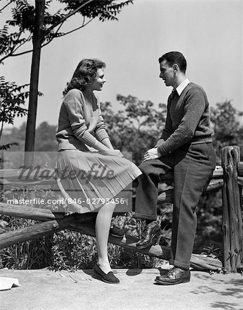1940s YOUNG TEENAGE COUPLE SITTING TALKING ON RUSTIC WOODEN FENCE