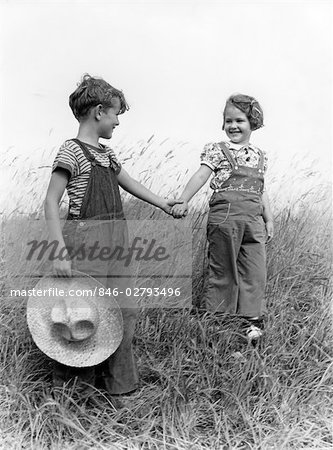 1930s 1940s FARM KIDS BOY GIRL WEARING OVERALLS HOLDING HANDS WALK DOWN GRASSY HILL BOY CARRYING STRAW HAT