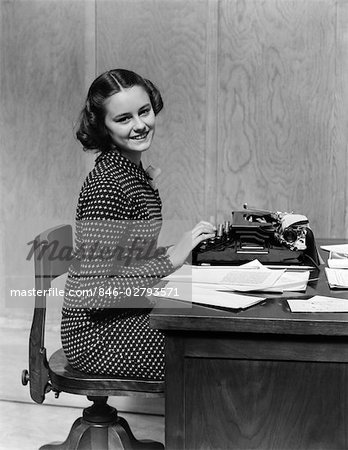 1940s YOUNG WOMAN OFFICE SECRETARY SMILING SITTING AT DESK USING MANUAL TYPEWRITER LOOKING AT CAMERA
