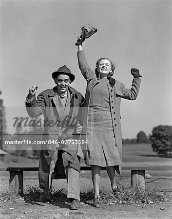 1940s YOUNG TEEN COUPLE BOY GIRL BENCH WEARING COATS CHEERING AT SPORTING EVENT EXCITED HAPPY SPECTATORS