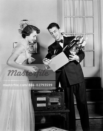 1950s TEEN COUPLE BOY AND GIRL IN PROM FORMAL WEAR PLAYING PHONOGRAPH RECORDS IN HOME LIVING ROOM