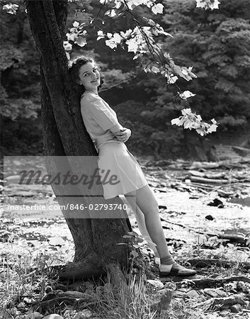 1940s SMILING YOUNG WOMAN WEARING SHORT SKIRT AND SADDLE SHOES LEANING AGAINST TREE BY STREAM