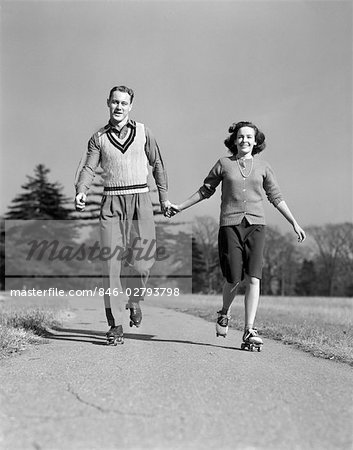 1940s SMILING TEENAGE COUPLE BOY GIRL HOLDING HANDS ROLLER SKATING ON SIDEWALK
