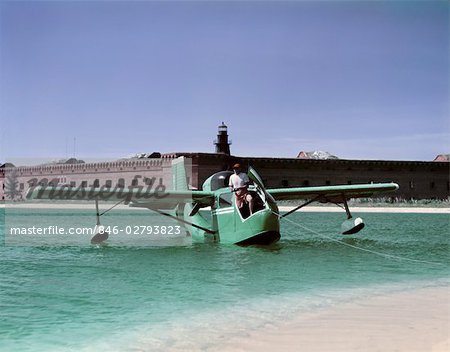 1950s MAN FISHING OFF FRONT OF SEAPLANE HYDROPLANE AIRCRAFT TURQUOISE BLUE GREEN WATER LIGHTHOUSE BRICK BUILDING HORIZON