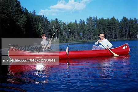 1960s GRANDFATHER & BOY RED CANOE ON LAKE FISHING GRANDSON REELING IN FISH CATCH FAMILY FAMILIES GENERATIONS