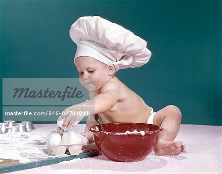 1960s BABY WEARING CHEFS HAT HOLDING EGG SURROUNDED BY COOKING EQUIPMENT
